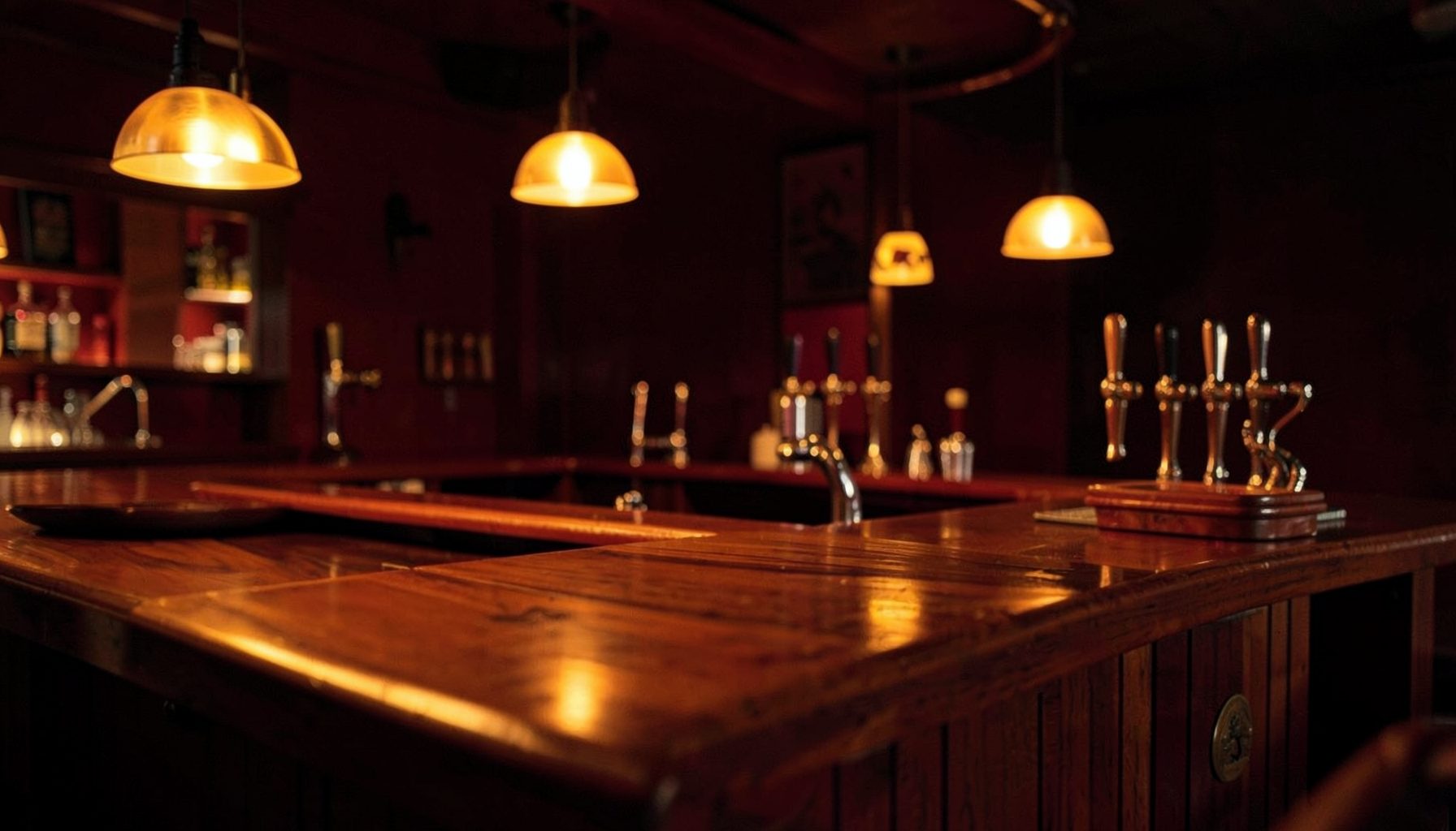 Warm amber and deep red bar interior with soft pendant lighting, polished wood surfaces, and a row of tap handles glowing